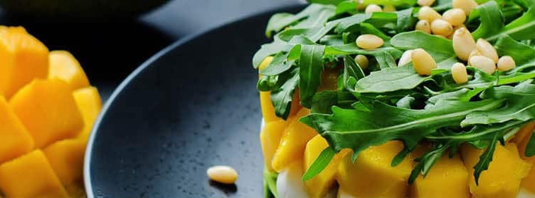 Close up of a mango-arugula dish with nuts on top on a black plate.