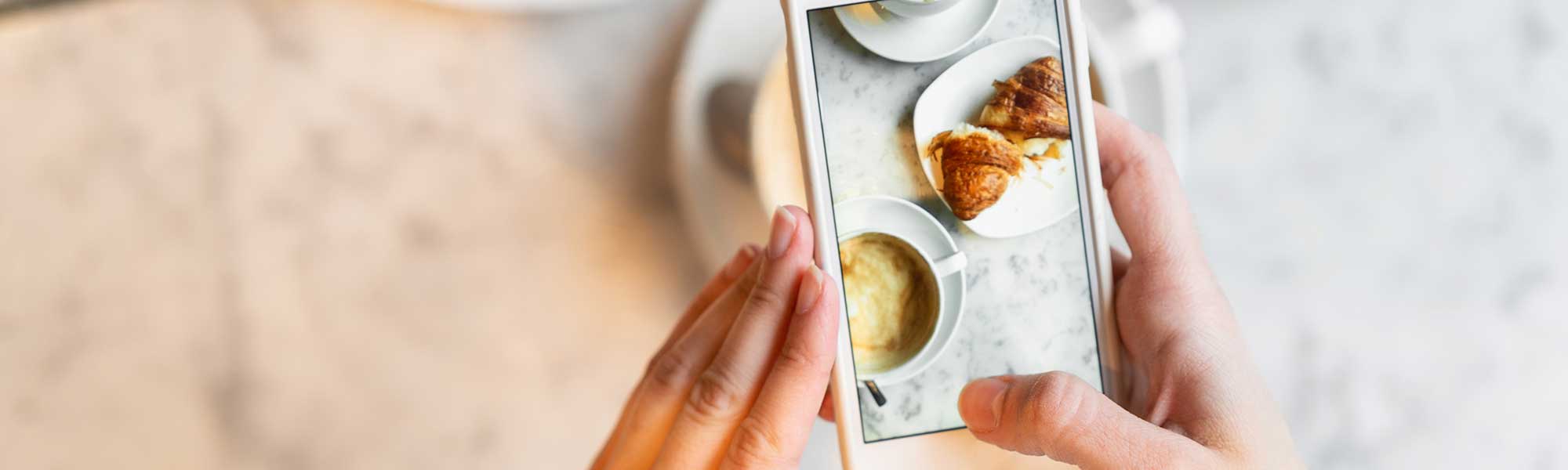 Hands taking photo of croissant and cup of coffee with a smartphone.