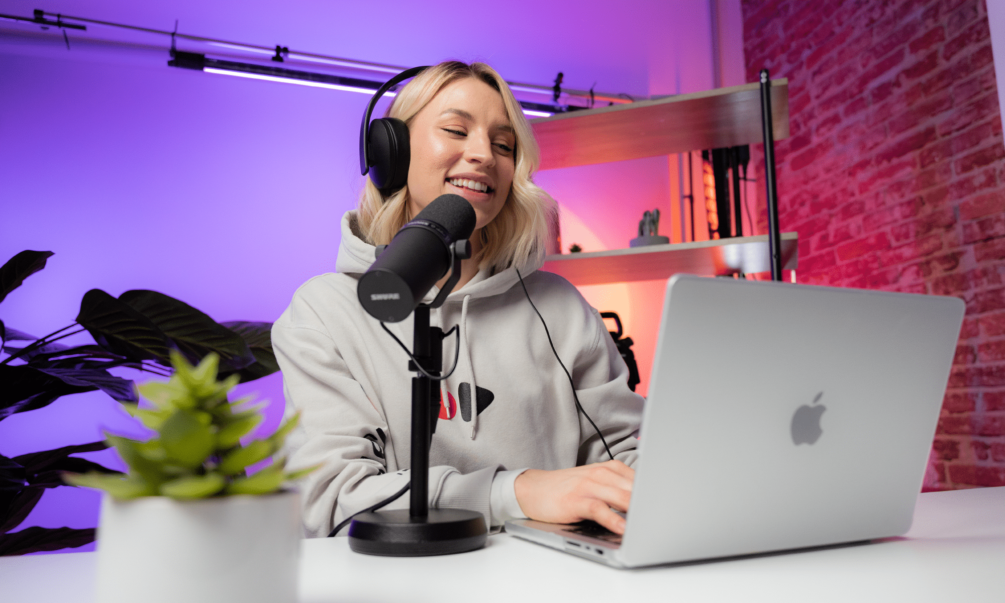 A woman smiles as she speaks into a microphone and records content into her Mac laptop.