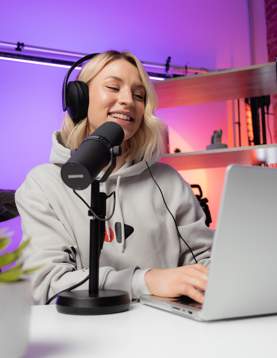 A woman smiles as she speaks into a microphone and records content into her Mac laptop.