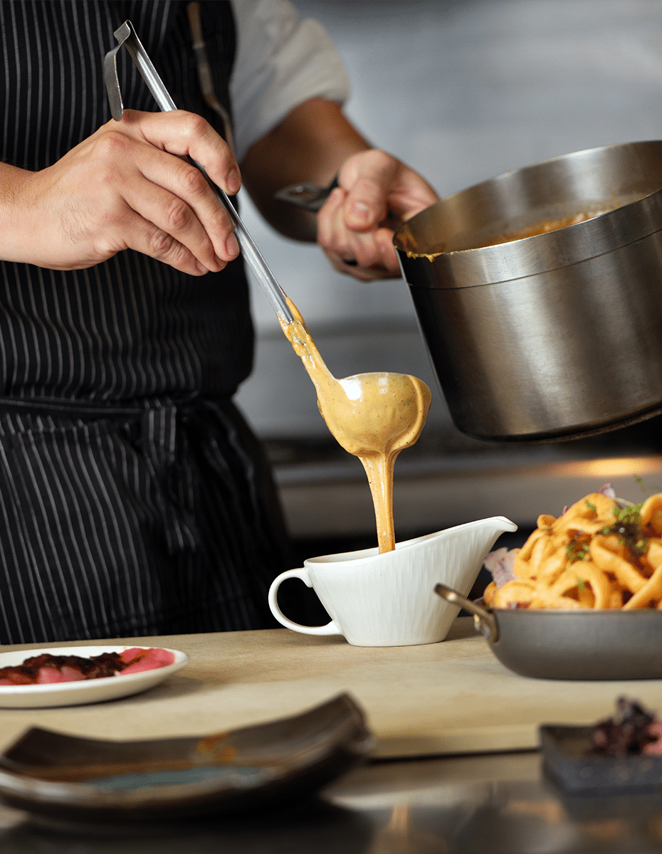 A chef pours hot cheese gravy into a white serving pourer next to an already-prepared dish.